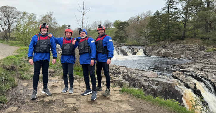 Group of men standing in wet suits looking at camera with low force waterfall in the background. 